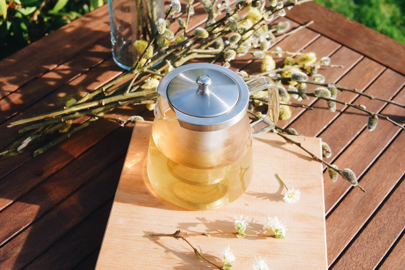 Skleněná konvice na čaj Simax Charme a jarní květiny položené na dřevěném zahradním stole s velikonočními kočičkami v pozadí
