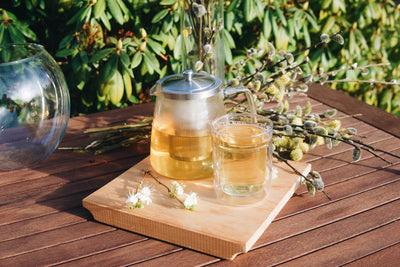 Skleněná konvice na čaj Simax Charme a skleněný hrnek Simax Twin na dřevěném zahradním stole s jarními květinami