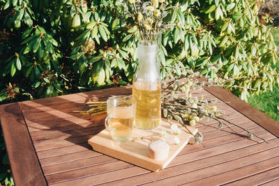Skleněná karafa Simax Bastia, sklenice Simax Lyra plné čaje a velikonoční kočičky položené na dřevěném zahradním stole