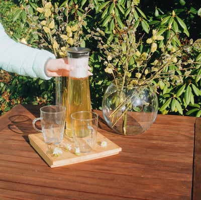 Skleněná karafa na čaj Simax Indis s čajem na zahradním stole. V popředí dvě sklenice na čaj Simax Lyra, v pozadí skleněné vázy Simax Globe a Drum