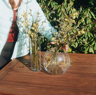 Kulatá skleněná váza Simax Globe a vysoká skleněná váza Simax Drum s velikonočními kočičkami na dřevěném zahradním stole