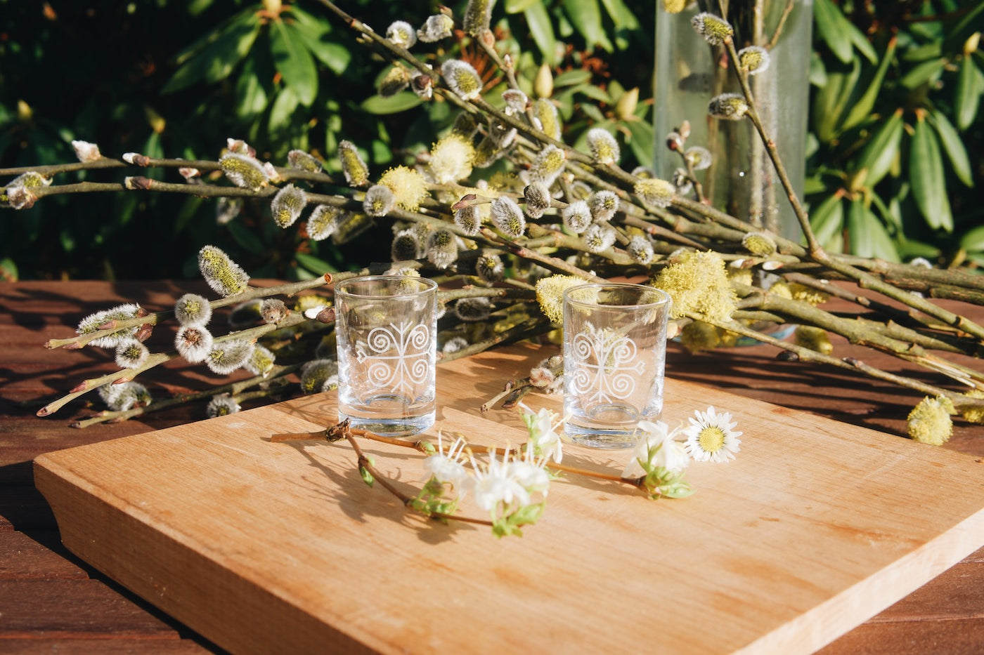 Skleněné štamprle na slivovici Ludové 30 ml Ateliér Žampach a jarní květiny položené na dřevěném zahradním stole. V pozadí velikonoční kočičky