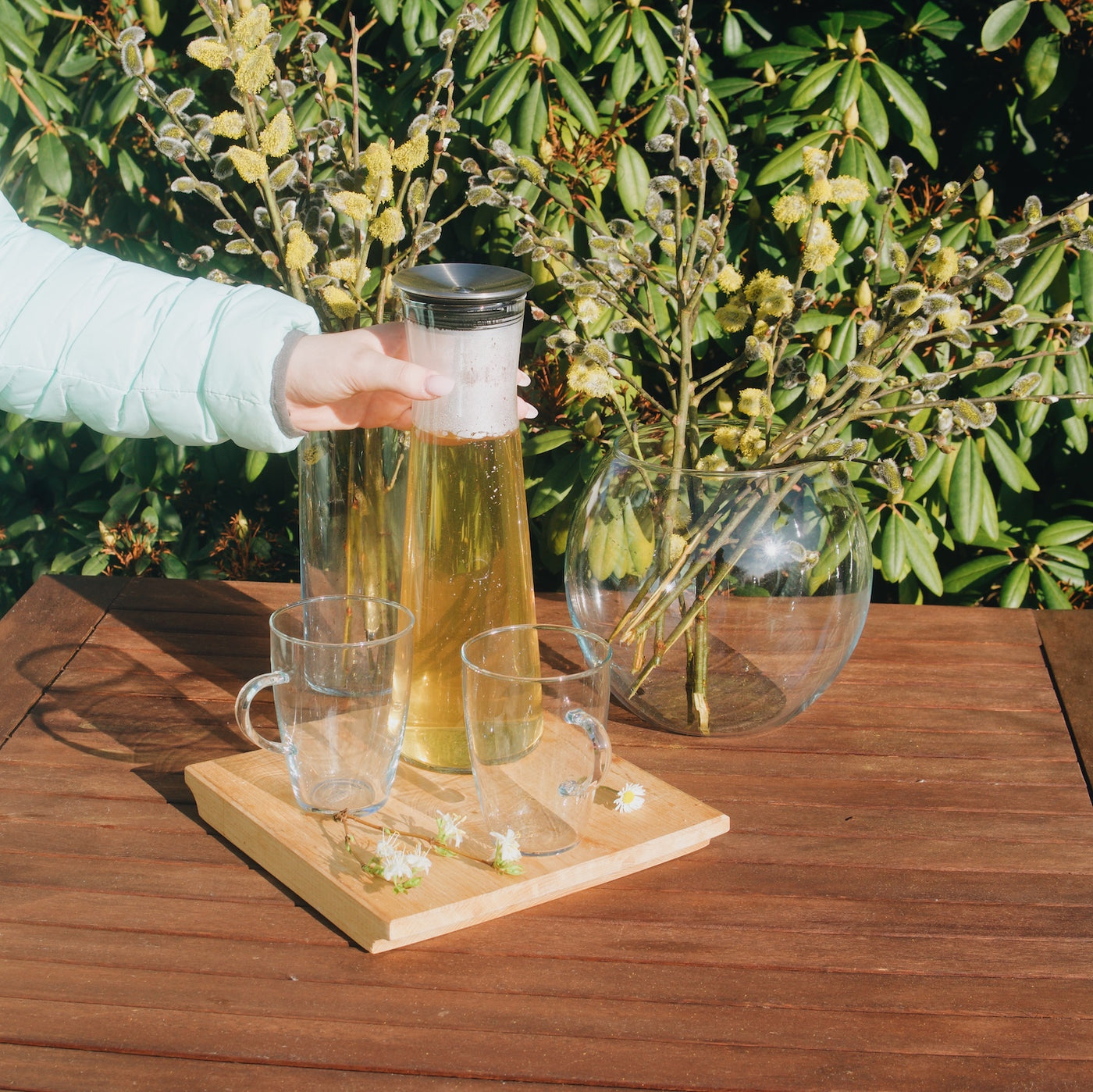 Dvě sklenice na čaj Simax Lyra a skleněná karafa Simax Indis na dřevěném zahradním stole. V pozadí skleněné vázy Simax Drum a Globe.